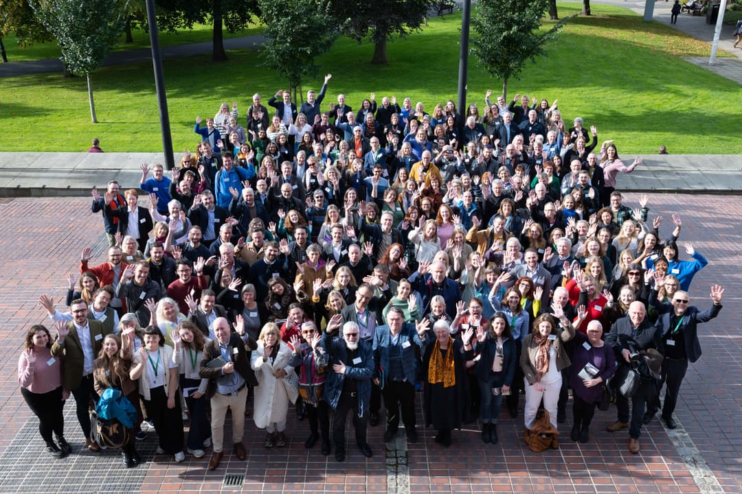 A large group of people stand outside and look up at the photographer. They are smiling and waving.